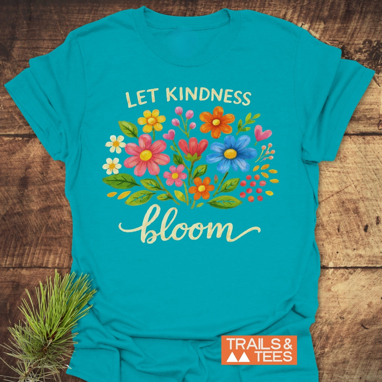 Let Kindness Bloom T-Shirt featuring a vibrant floral bouquet and slogan, displayed on a wooden surface, embodies durable ring-spun cotton comfort ideal for outdoor adventures and everyday wear.