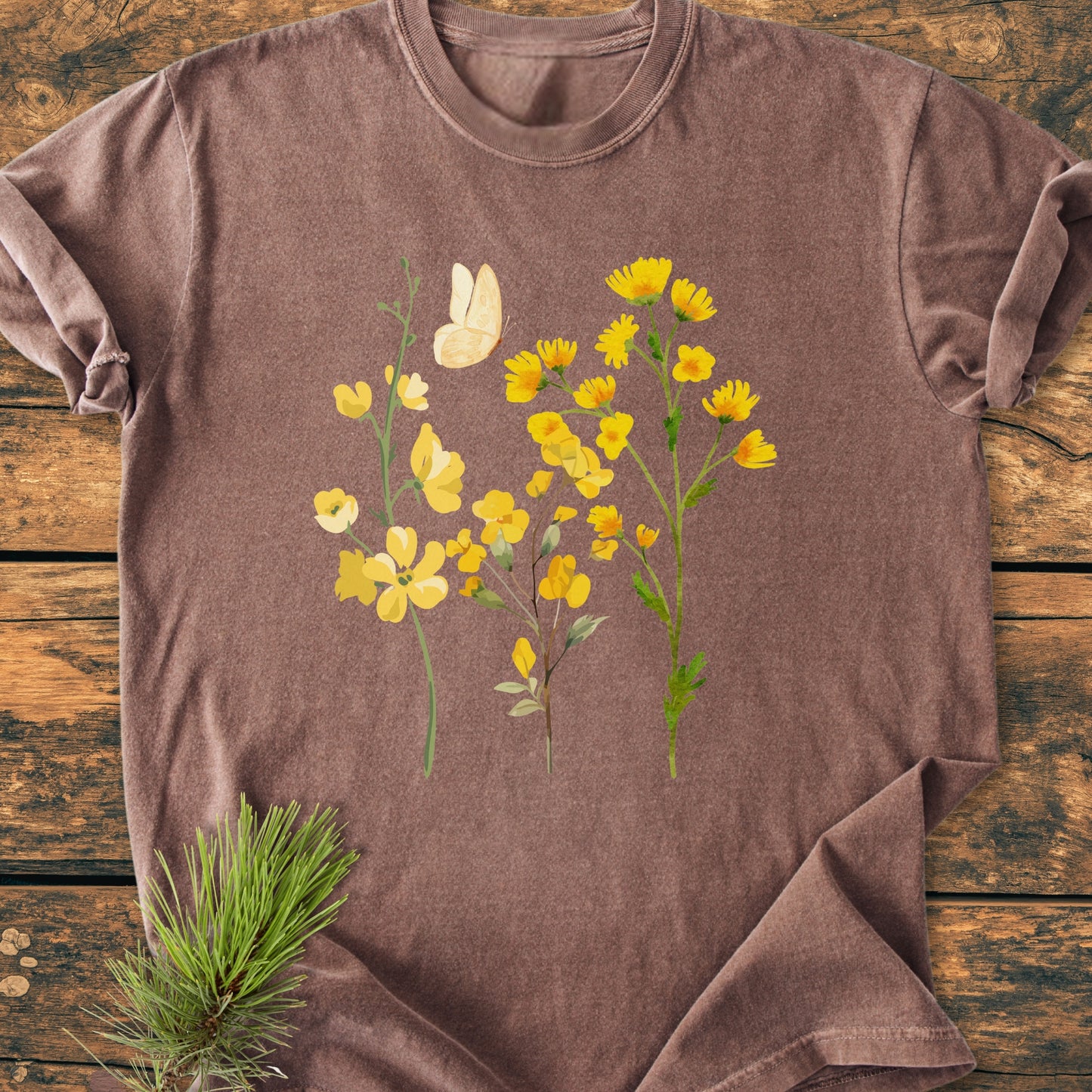 The Yellow Boho Flowers - Vintage Heavyweight Tee features stylized yellow wildflowers and a cream butterfly on a heather-brown fabric with rolled sleeves, laid on rustic wood.