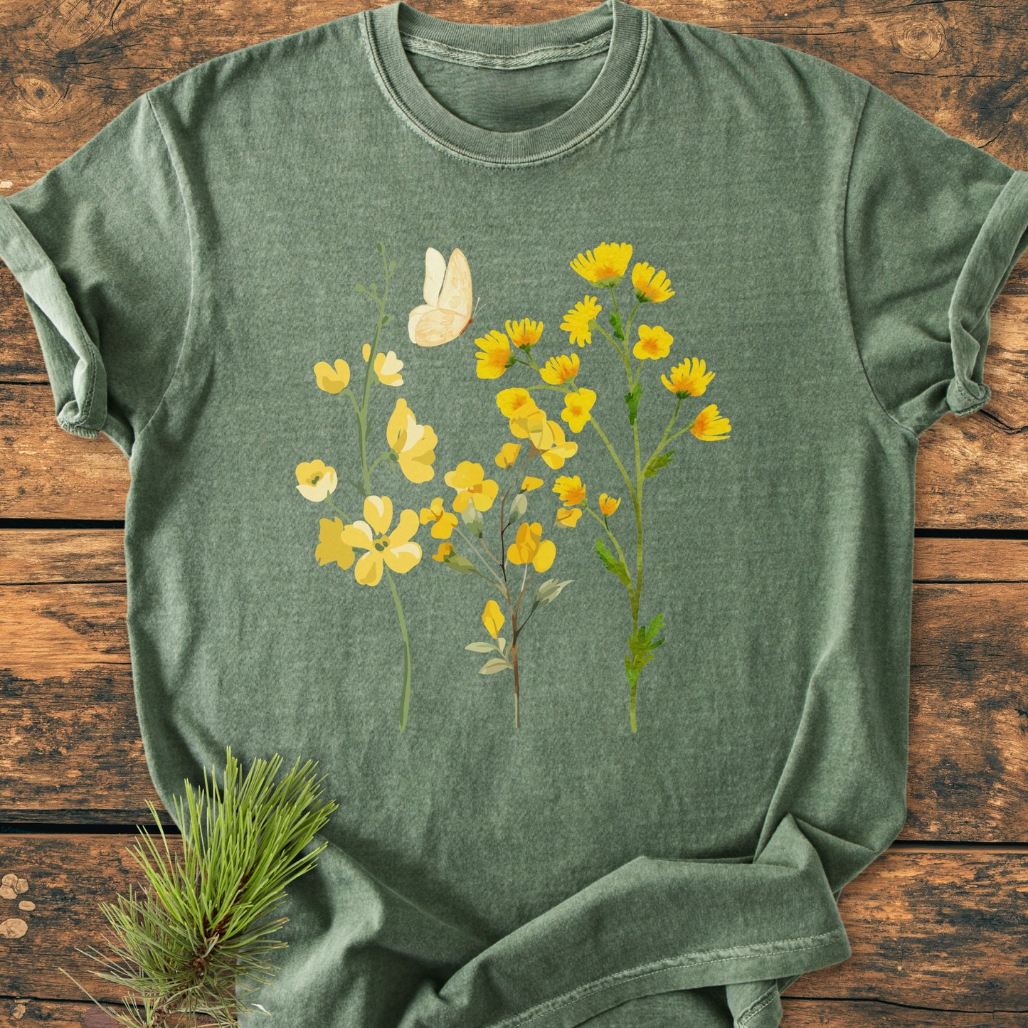 Yellow Boho Flowers - Vintage Heavyweight Tee features a botanical graphic with wildflowers and butterfly on a heather-green T-shirt, styled on a wooden surface with a pine branch accent.