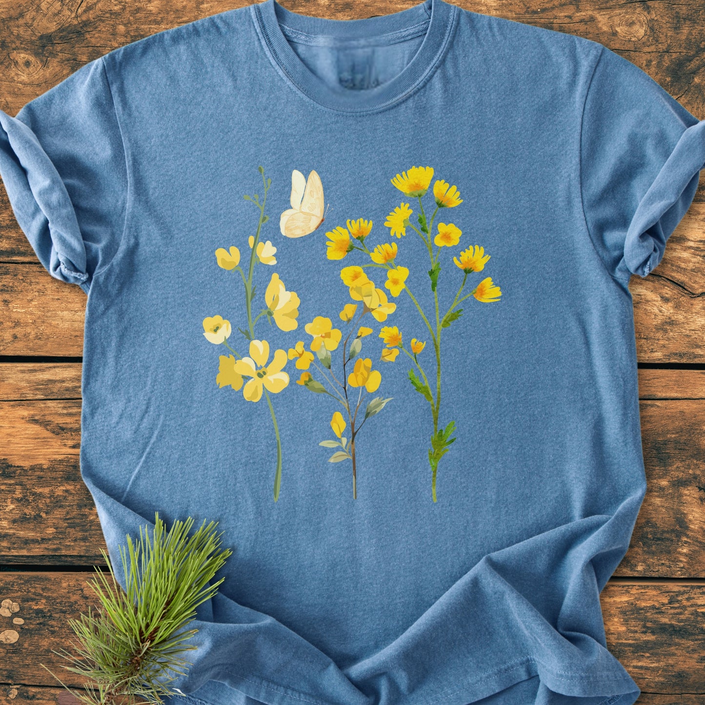Yellow Boho Flowers - Vintage Heavyweight Tee, showcasing bright yellow wildflowers and a butterfly on a blue cotton fabric, laid on a wooden surface with a pine sprig accent.