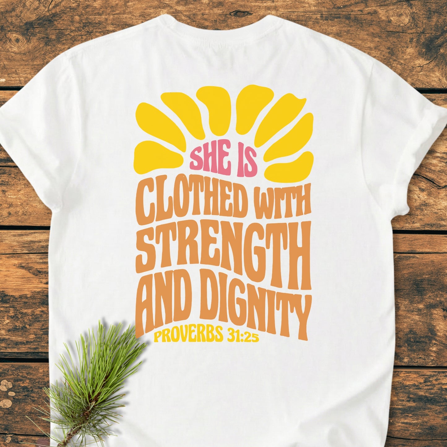 Strength And Dignity BACK PRINT - Vintage Wash Tee: White T-shirt with a yellow sun graphic and inspirational text on the back, displayed flat with a pine sprig on wooden surface.