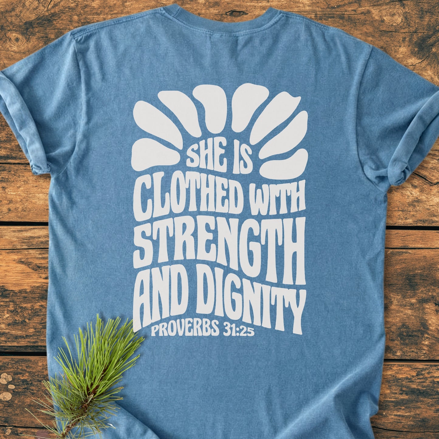 Strength And Dignity BACK PRINT - Vintage Wash Tee, featuring Proverbs 31:25 in bold white on soft-blue, laid flat on wood with pine sprig; a garment-dyed, relaxed fit, 100% ring-spun cotton T-shirt.