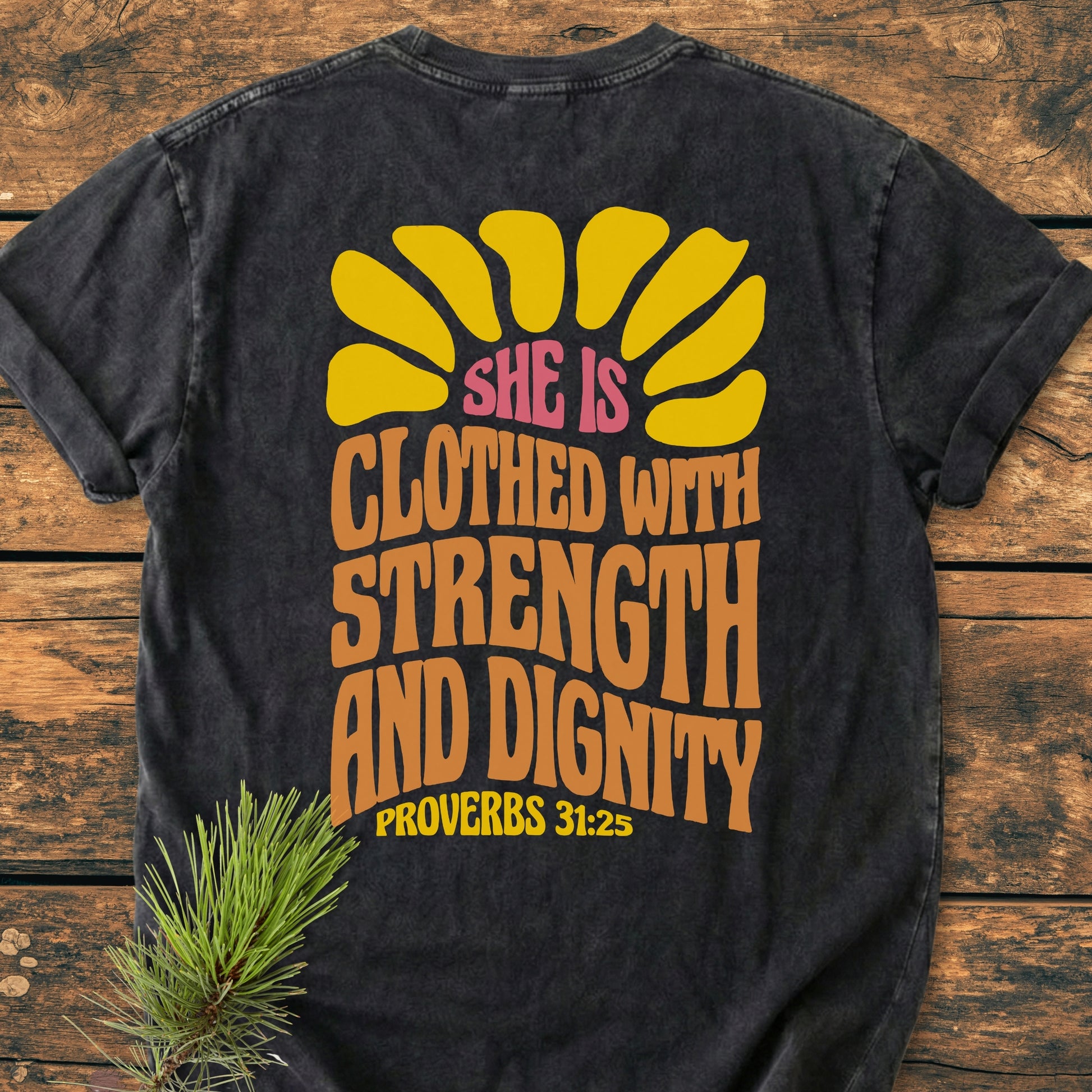 Strength And Dignity BACK PRINT - Vintage Wash Tee features a retro yellow sunburst and Proverbs 31:25 on dark-charcoal fabric, laid on wood with a pine branch accent.