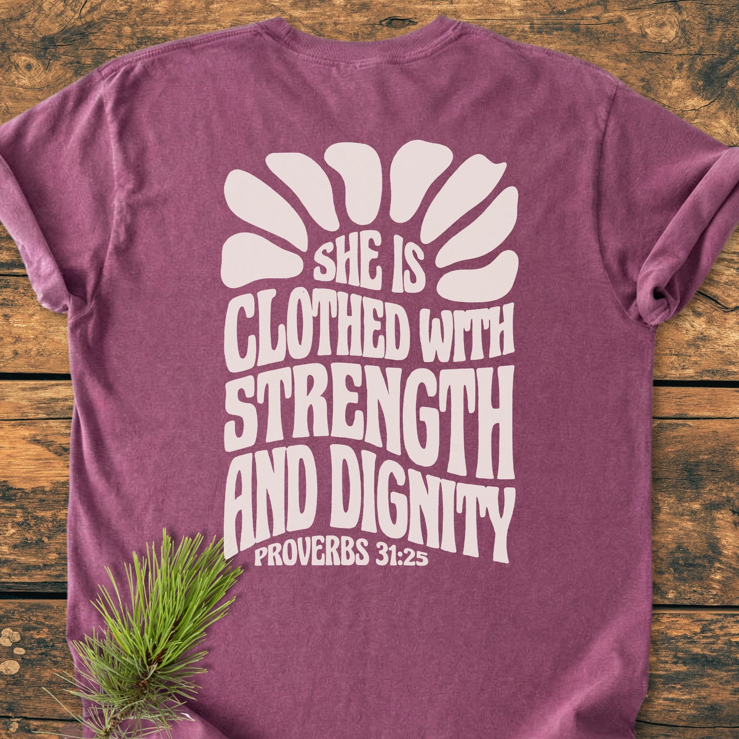 Strength And Dignity BACK PRINT - Vintage Wash Tee: Dusty-rose T-shirt with white rising-sun graphic and Proverbs 31:25 text on the back, pine sprig accent, laid on wooden tabletop.