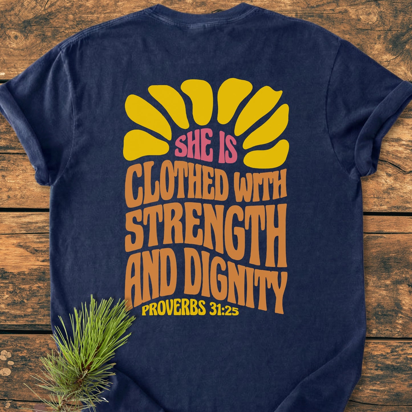 Strength And Dignity BACK PRINT - Vintage Wash Tee: Navy-blue tee with mustard half-sun and Proverbs 31:25 quote on back. Laid flat on rustic wood with a pine sprig beside.