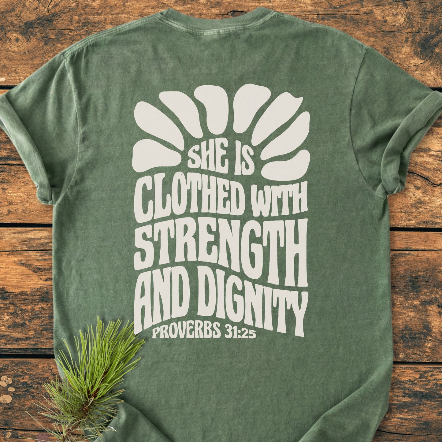 Strength And Dignity BACK PRINT - Vintage Wash Tee: Sage-green T-shirt with short sleeves, featuring a white sunburst graphic and Proverbs 31:25 quote on the back, laid flat on a wooden surface.