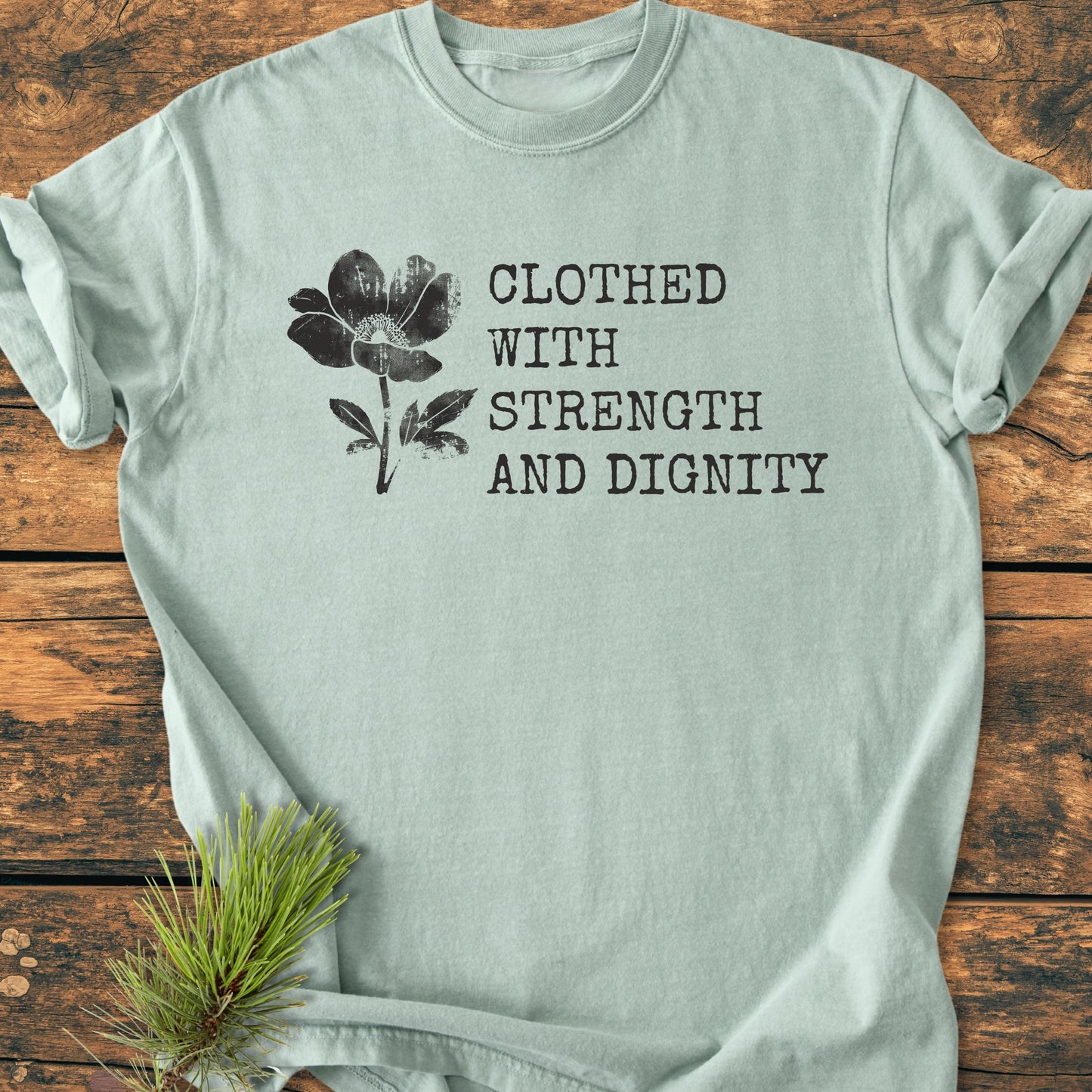 Clothed With Strength And Dignity, Proverbs 31:25 - Vintage Heavyweight Tee featuring a distressed black flower graphic and empowering phrase on a soft, pale-green crew-neck T-shirt, styled on rustic wood.