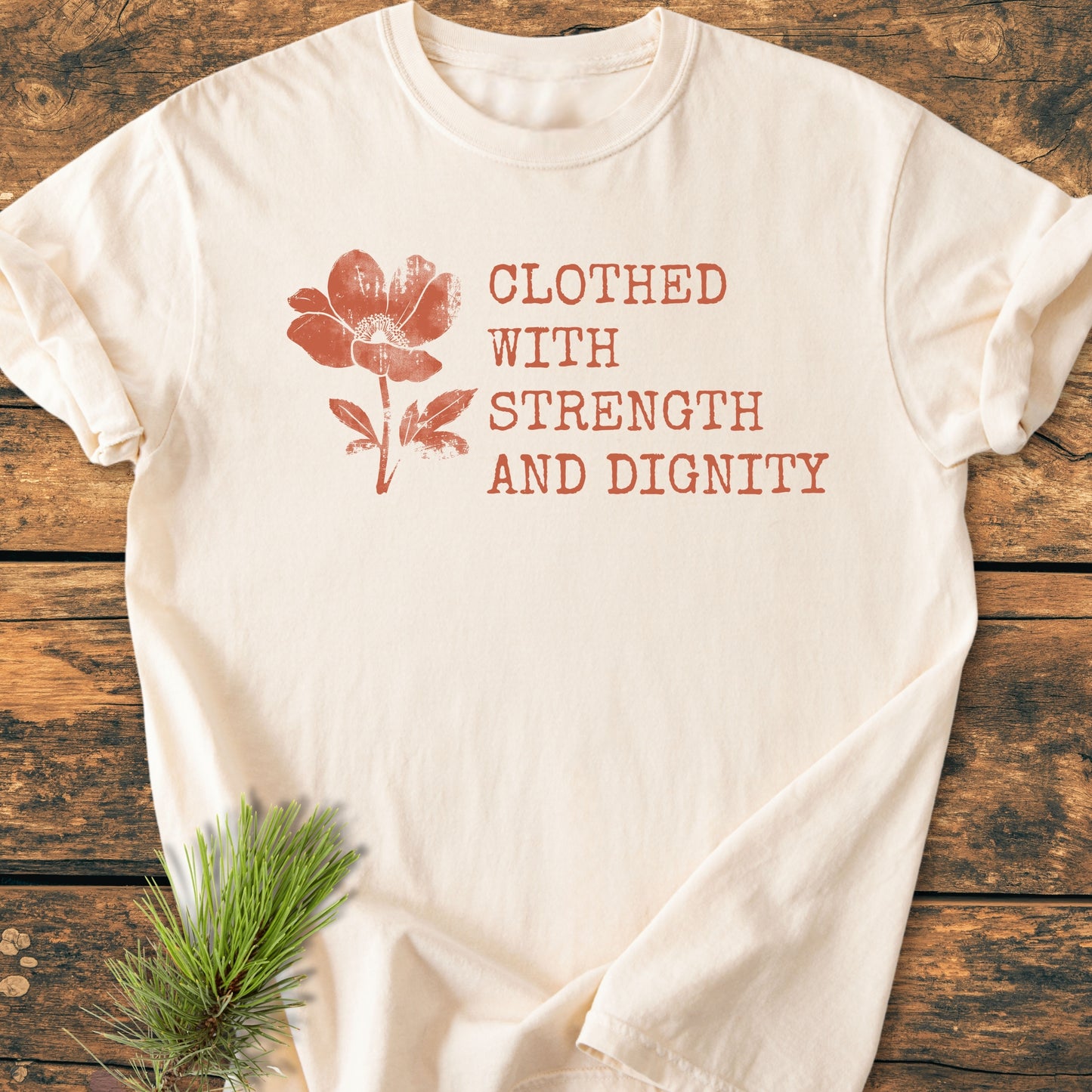 Clothed With Strength And Dignity, Proverbs 31:25 - Vintage Heavyweight Tee with a distressed blossom illustration and rustic font, laid flat on wood, accented by an evergreen sprig.