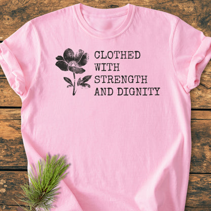 Clothed With Strength And Dignity, Proverbs 31:25 - Vintage Heavyweight Tee features a black flower graphic and typewriter text on soft pink, laid flat on a rustic wooden surface with pine needles.