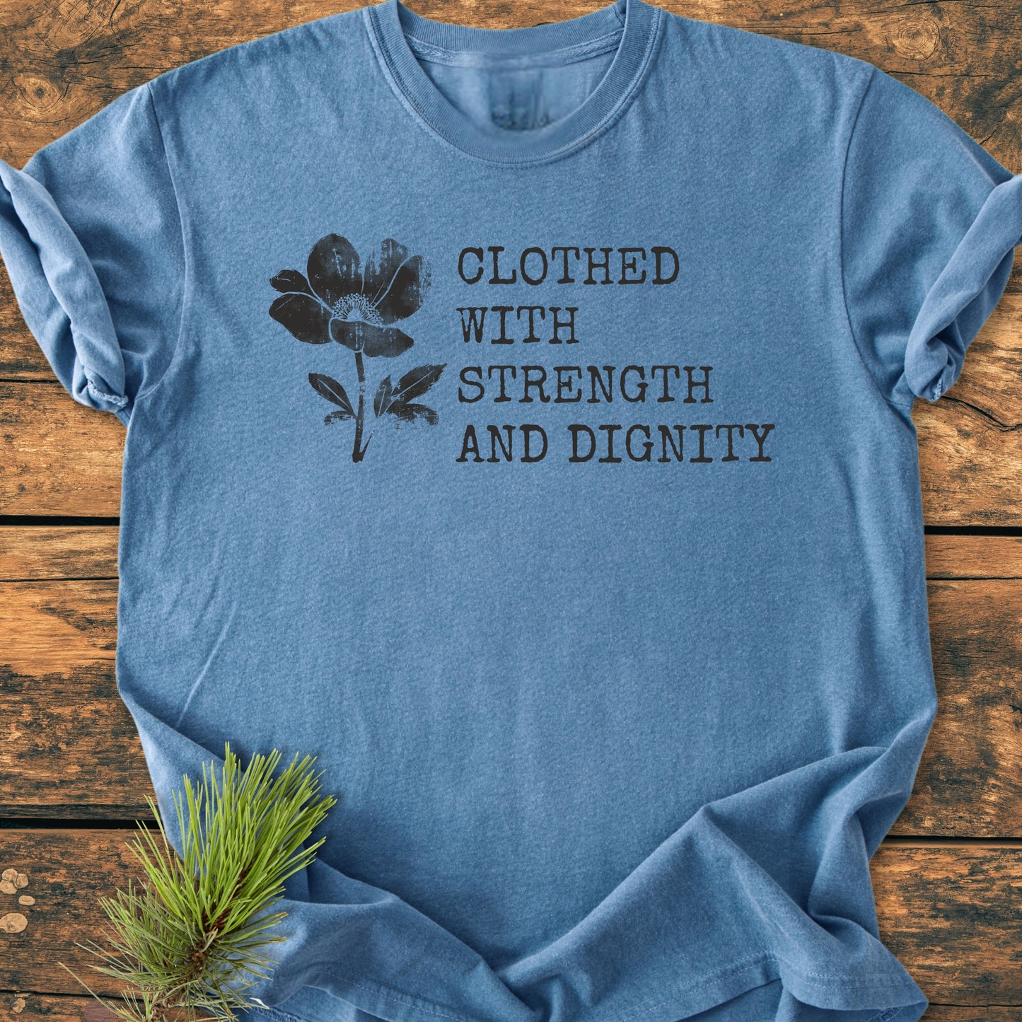 Heather-blue crew-neck tee, Clothed With Strength And Dignity, Proverbs 31:25 in distressed black font, floral illustration, laid on wood surface with pine sprig.