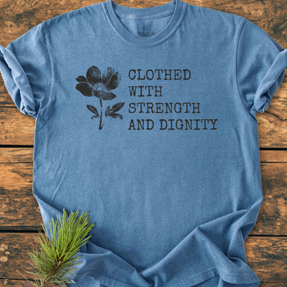 Heather-blue crew-neck tee, Clothed With Strength And Dignity, Proverbs 31:25 in distressed black font, floral illustration, laid on wood surface with pine sprig.