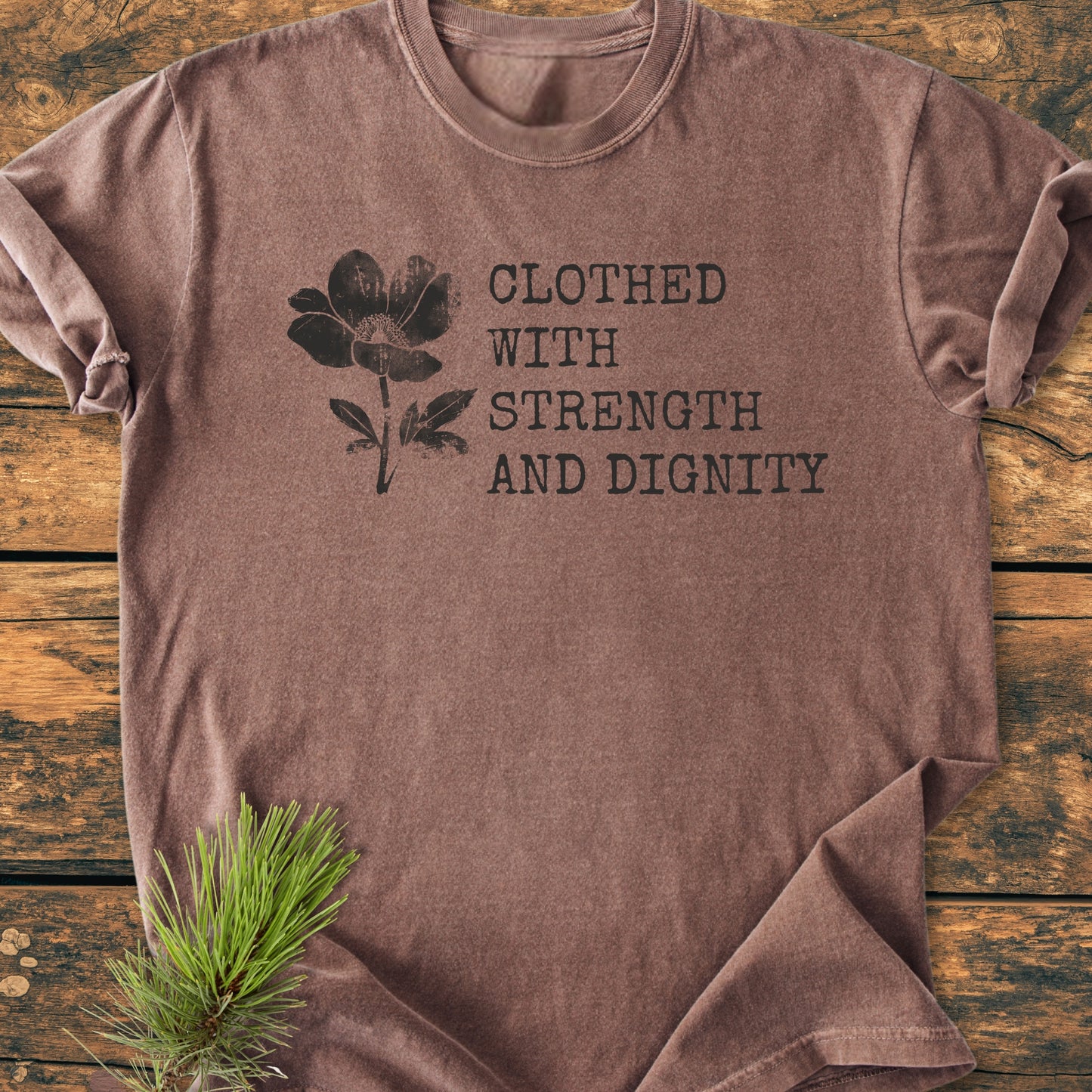 Clothed With Strength And Dignity, Proverbs 31:25 - Vintage Heavyweight Tee features a flower silhouette and quote on a brown T-shirt, displayed on wood with a pine sprig accent.