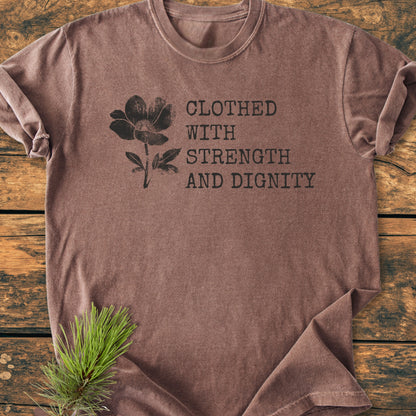 Clothed With Strength And Dignity, Proverbs 31:25 - Vintage Heavyweight Tee features a flower silhouette and quote on a brown T-shirt, displayed on wood with a pine sprig accent.