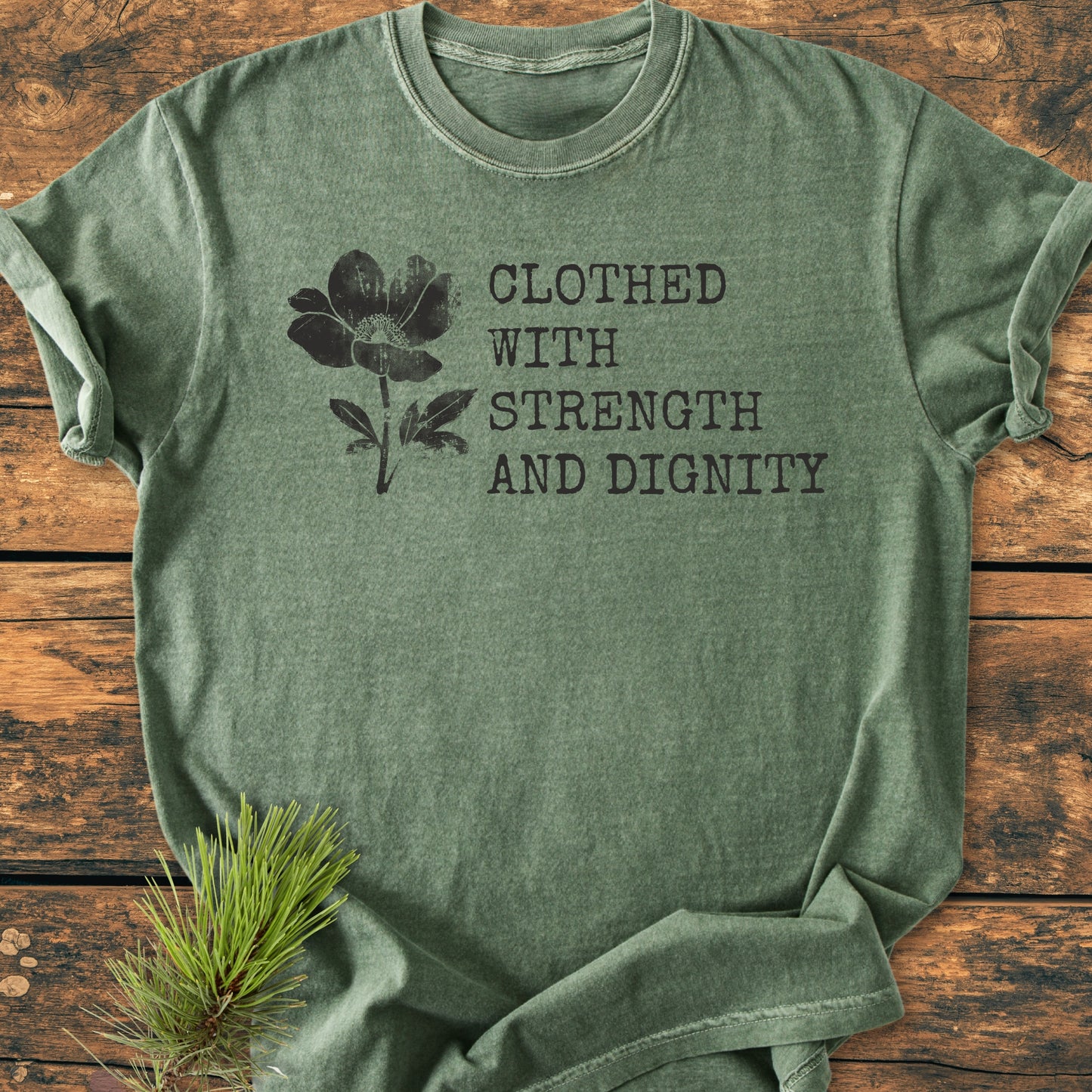 Heather-green T-shirt with Clothed With Strength And Dignity, Proverbs 31:25 in bold letters, featuring a flower silhouette, laid on wood with a pine sprig, emphasizing a natural, rustic vibe.
