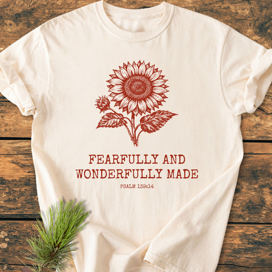 Fearfully And Wonderfully Made, Psalm 139:14 - Vintage Wash Tee features a stylized sunflower graphic and text on a light-colored, soft-washed T-shirt, laid on wood, with a pine sprig accent.