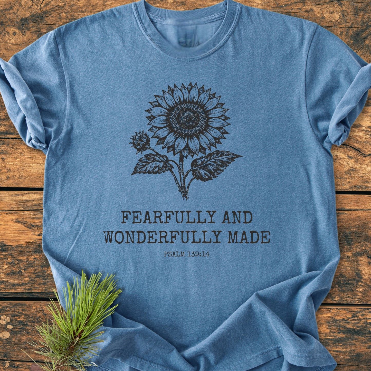 Washed-blue T-shirt with sunflower graphic, Fearfully And Wonderfully Made, Psalm 139:14, laid on wood surface, showcasing soft-washed texture, relaxed fit, and durable craftsmanship, enhanced by a pine sprig accent.
