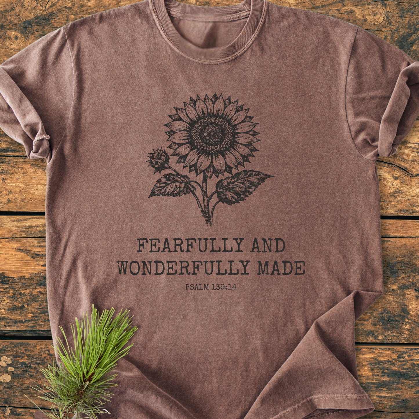 Fearfully And Wonderfully Made, Psalm 139:14 - Vintage Wash Tee features a sunflower line-drawing and text on a soft, brown vintage-washed T-shirt, laid on rustic wood with a pine sprig accent.