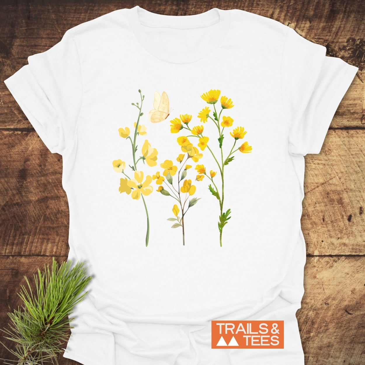 Yellow Boho Flowers T-Shirt featuring a trio of yellow wildflowers with a butterfly design, laid on rustic wood. Accentuated by a pine bough for a natural, outdoor vibe.