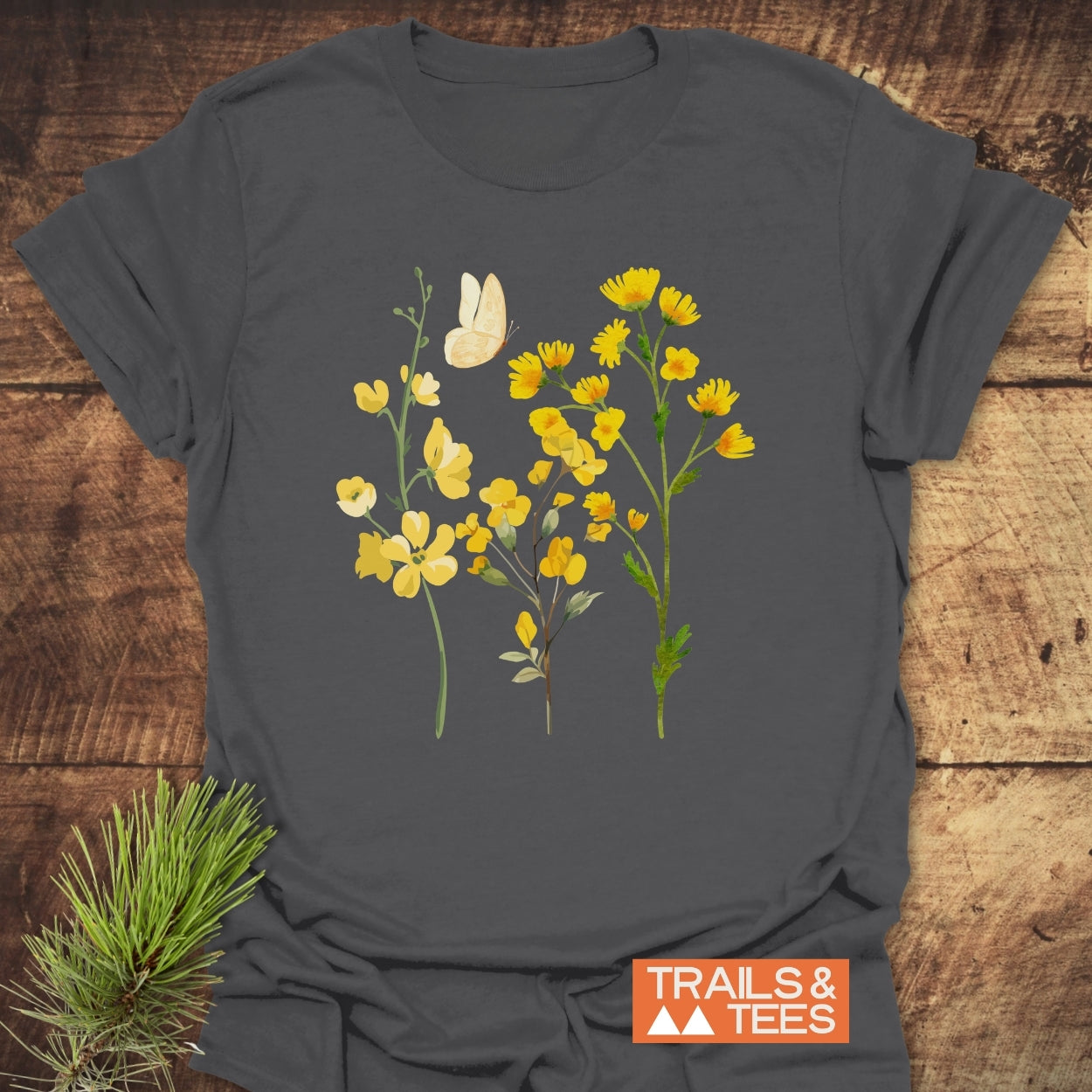 Yellow Boho Flowers T-Shirt featuring a botanical illustration of three yellow wildflower stems and a butterfly, laid on wood with a pine sprig, alongside TRAILS & TEES logo.
