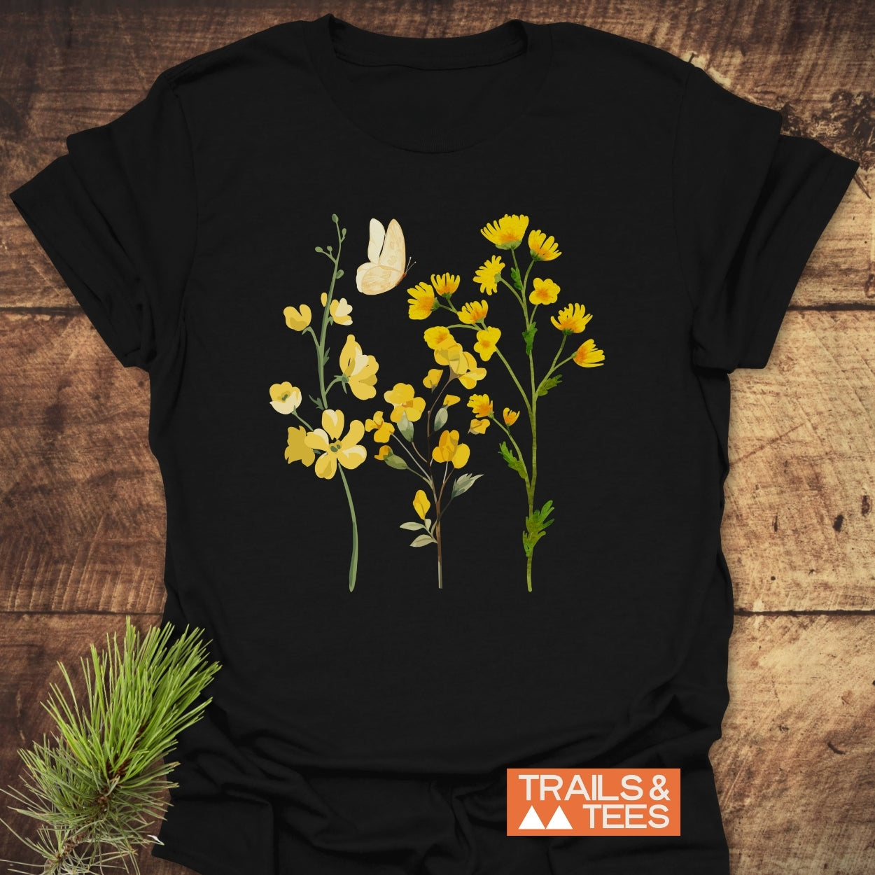 Yellow Boho Flowers T-Shirt featuring stylized yellow wildflowers and a butterfly on soft black cotton, with a “TRAILS & TEES” logo, laid out on a wooden surface.
