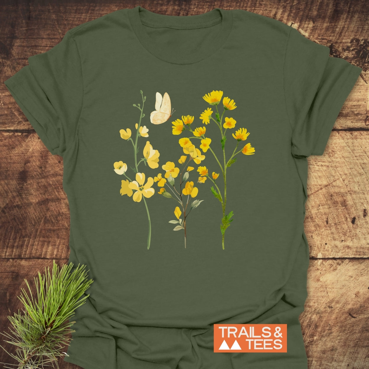 Yellow Boho Flowers T-Shirt featuring a botanical illustration of yellow wildflowers and a butterfly, laid on a wooden surface. Includes TRAILS & TEES logo, embodying outdoor adventure and comfort.