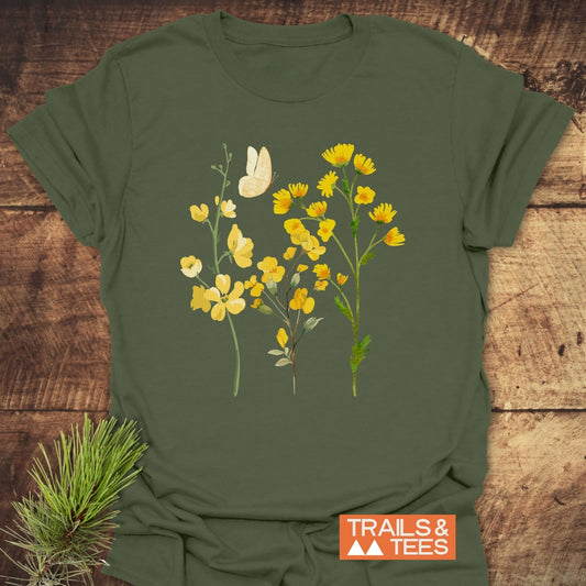 Yellow Boho Flowers T-Shirt featuring a botanical illustration of yellow wildflowers and a butterfly, laid on a wooden surface. Includes TRAILS & TEES logo, embodying outdoor adventure and comfort.