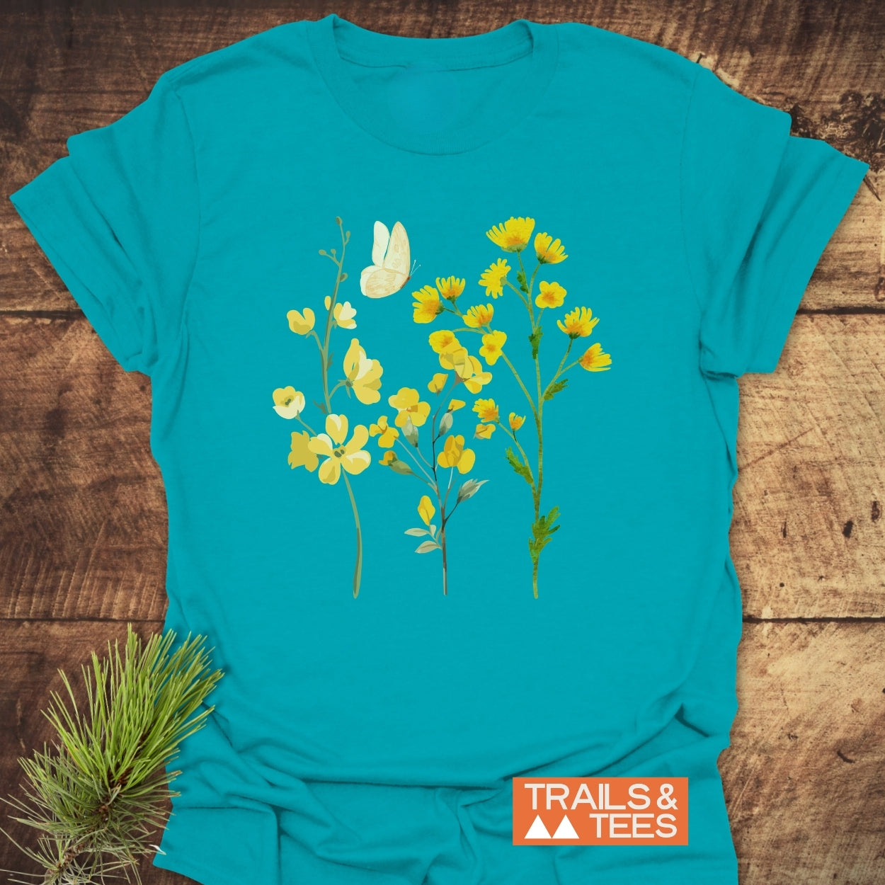 Yellow Boho Flowers T-Shirt with three stylized wildflowers and a butterfly on teal fabric, showcasing a nature-inspired design. A pine sprig complements the Trails & Tees logo beside the shirt.