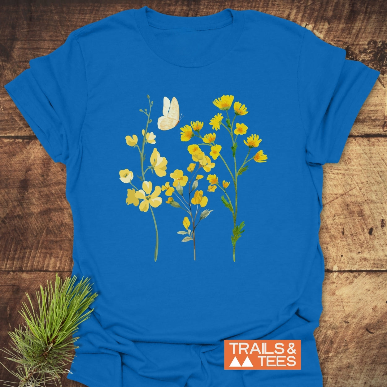 Yellow Boho Flowers T-Shirt featuring a botanical illustration of wildflowers and a butterfly, laid flat on wood. Includes TRAILS & TEES logo, pine needles tucked underneath.