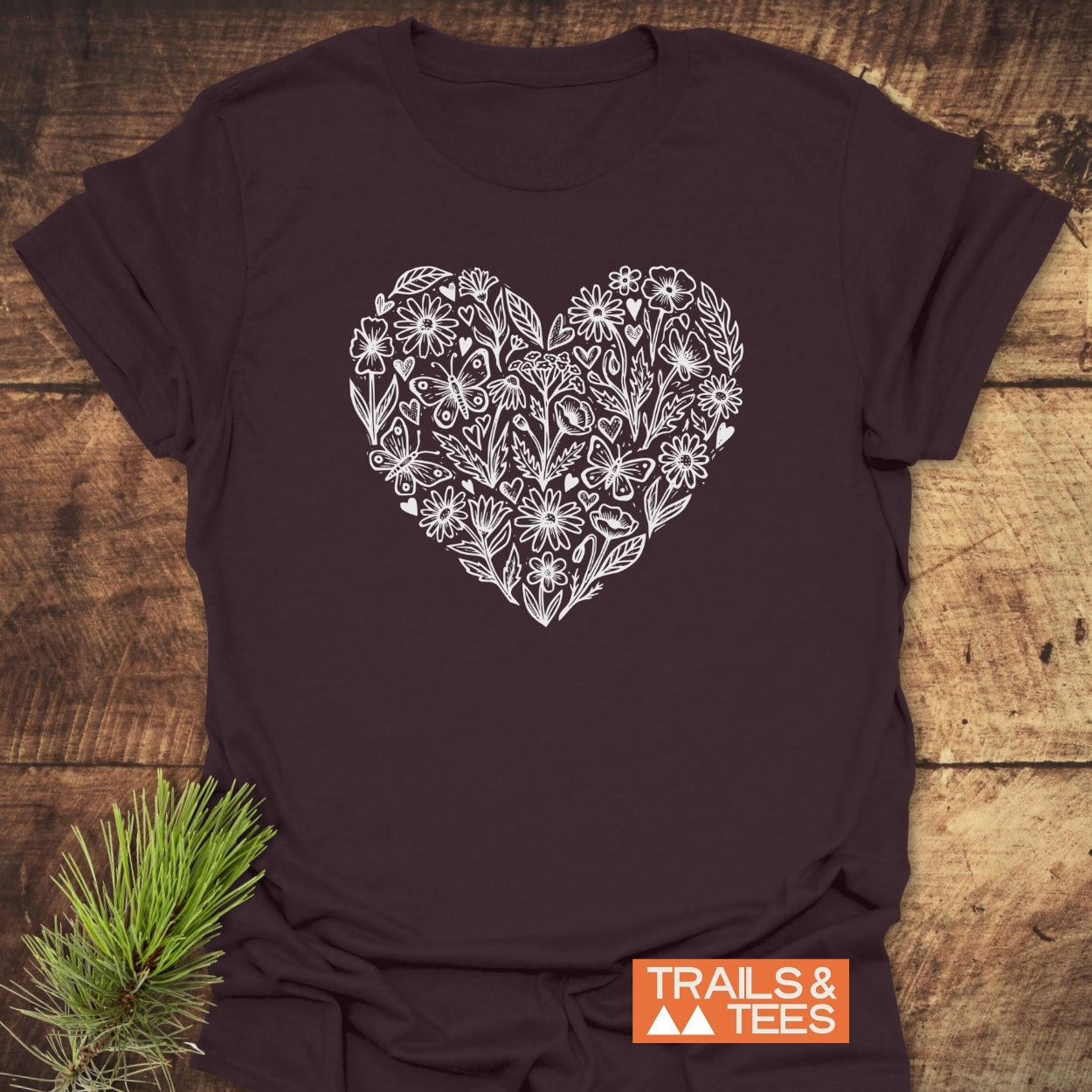 Wildflower Heart Butterfly Soul Softstyle T-Shirt featuring a white heart with sketched flowers, leaves, and butterflies, on a rustic wooden backdrop, highlighting TRAILS & TEES branding and pine needles.