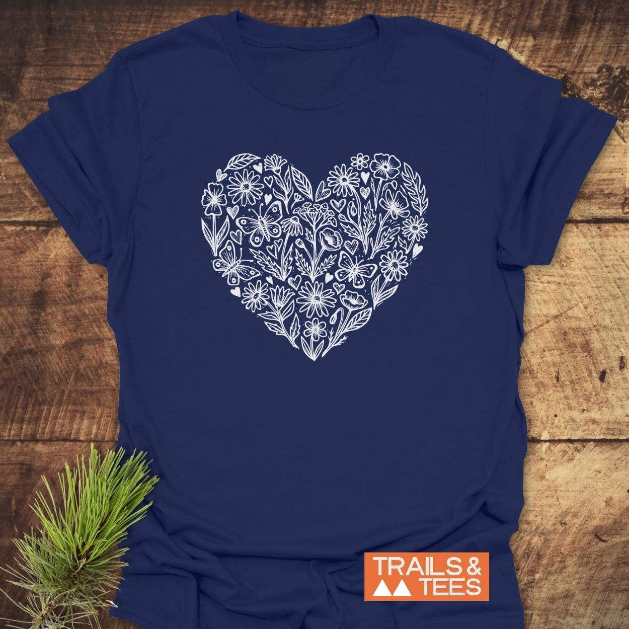 Wildflower Heart Butterfly Soul Softstyle T-Shirt with floral heart design on chest, laid on wood surface, showcases TRAILS & TEES logo. Pine needles accent for an outdoor-inspired look.