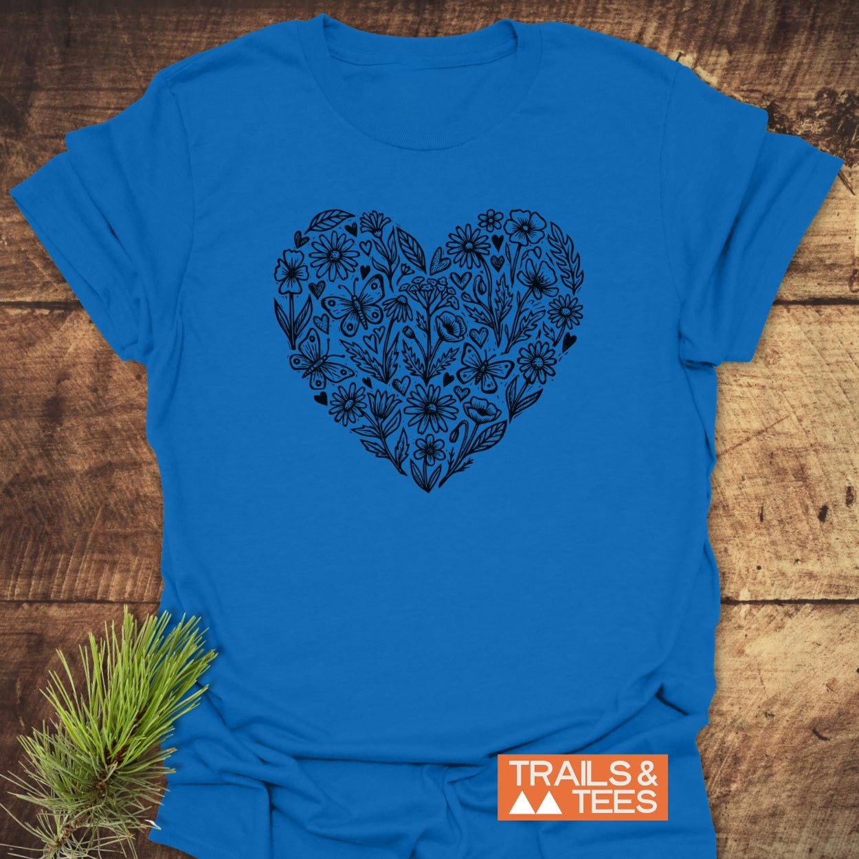 Wildflower Heart Butterfly Soul Softstyle T-Shirt featuring a black floral heart design on bright blue fabric, laid flat with a pine sprig. Includes an orange TRAILS & TEES label on the corner.