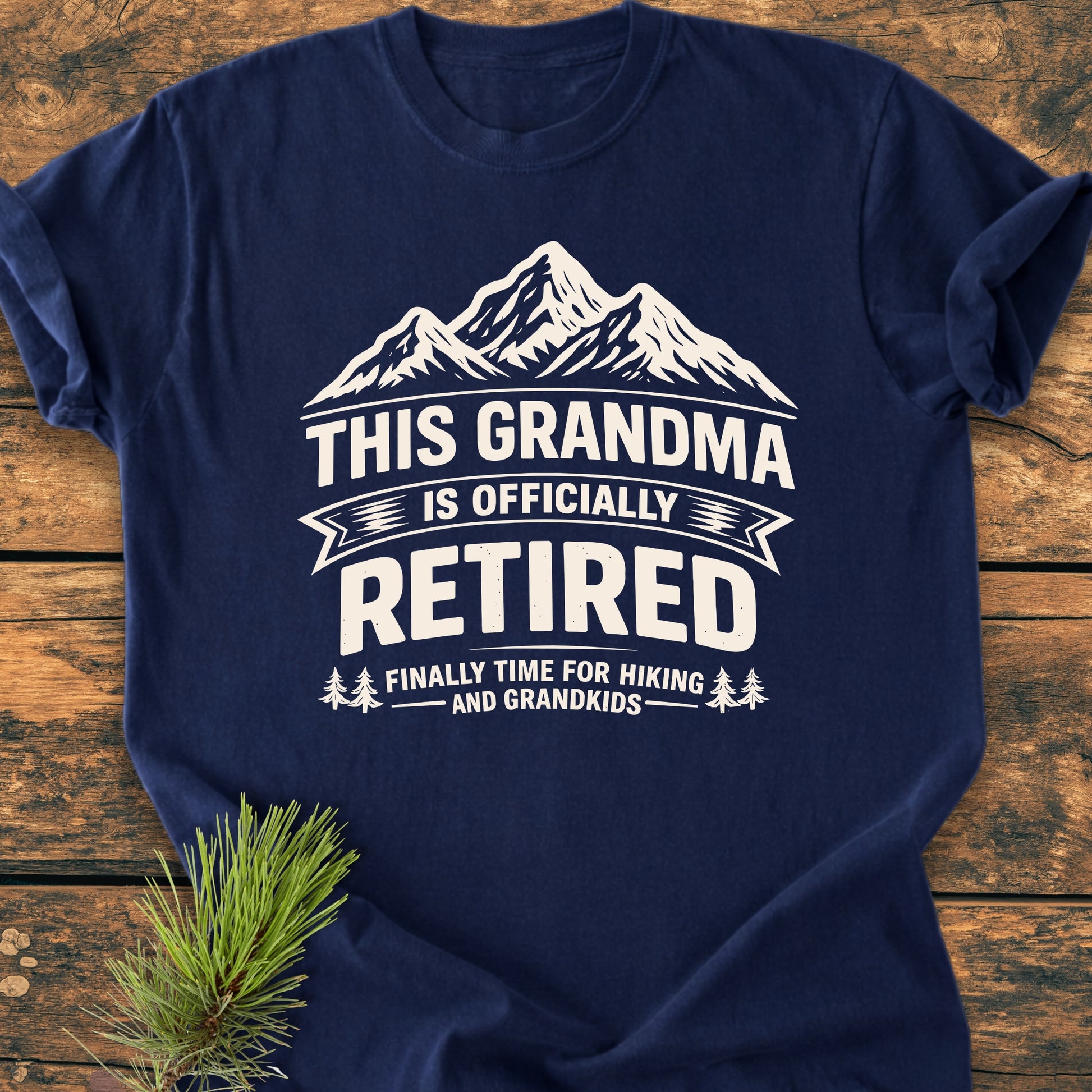 This Grandma is Officially Retired - Vintage Wash Tee features a mountain graphic and retirement slogan on a navy-blue T-shirt, laid on wood with a pine sprig, highlighting its cozy, relaxed fit.