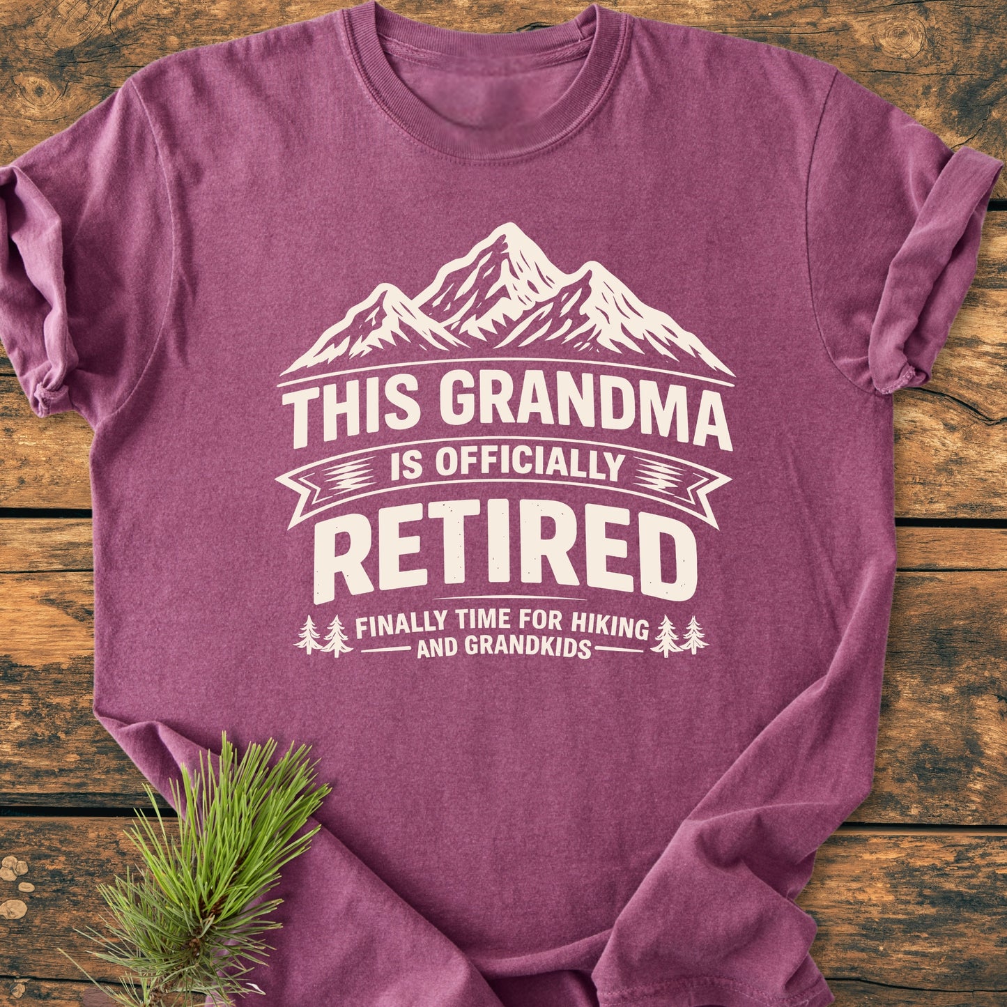 The This Grandma is Officially Retired - Vintage Wash Tee features a white mountain graphic with bold text on a heather-purple T-shirt, complemented by small tree icons and a pine sprig accent.