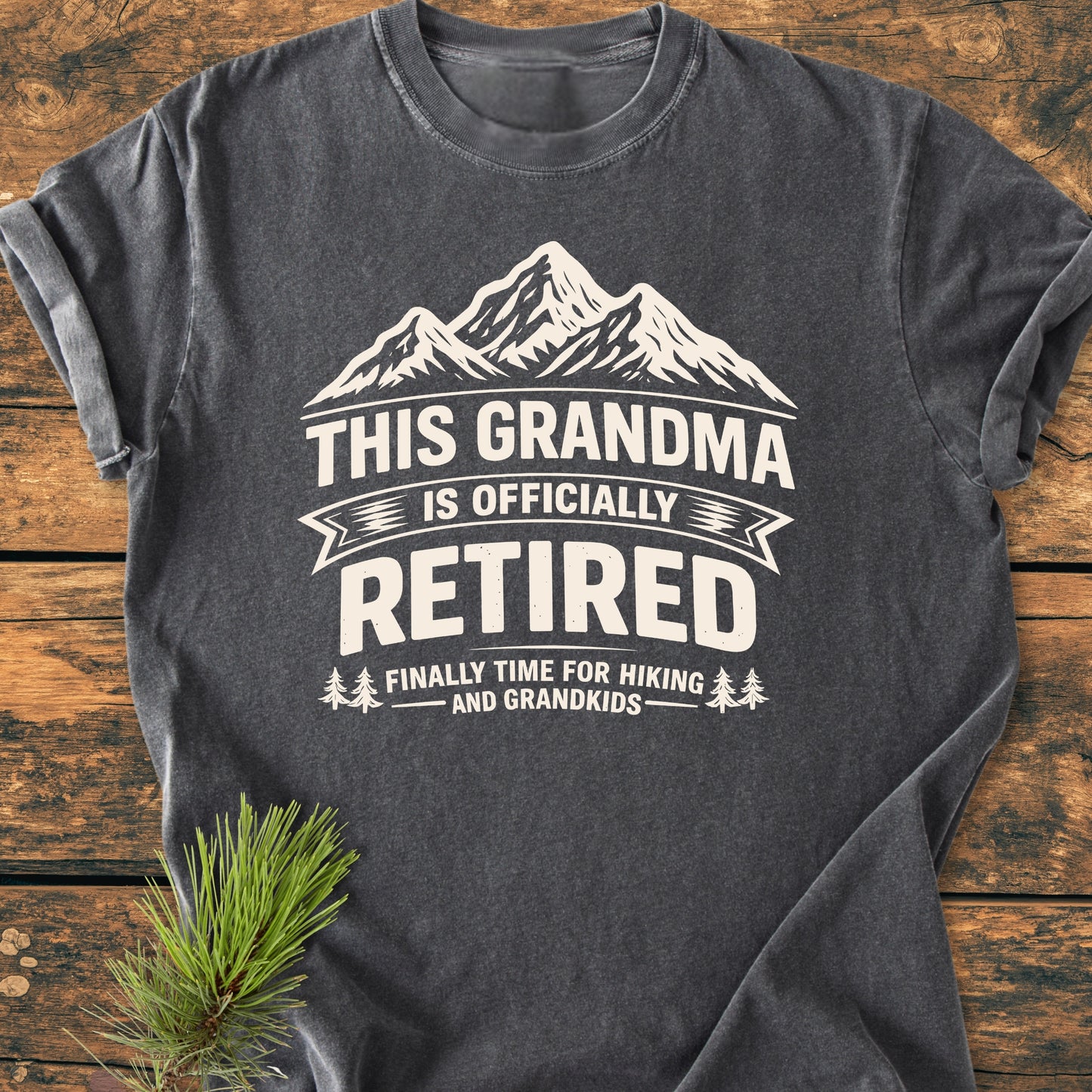 Charcoal-gray This Grandma is Officially Retired - Vintage Wash Tee features a white mountain-top graphic and slogan, laid on wood with pine sprig. Soft, cozy garment-dyed cotton, ideal for hiking and family time.
