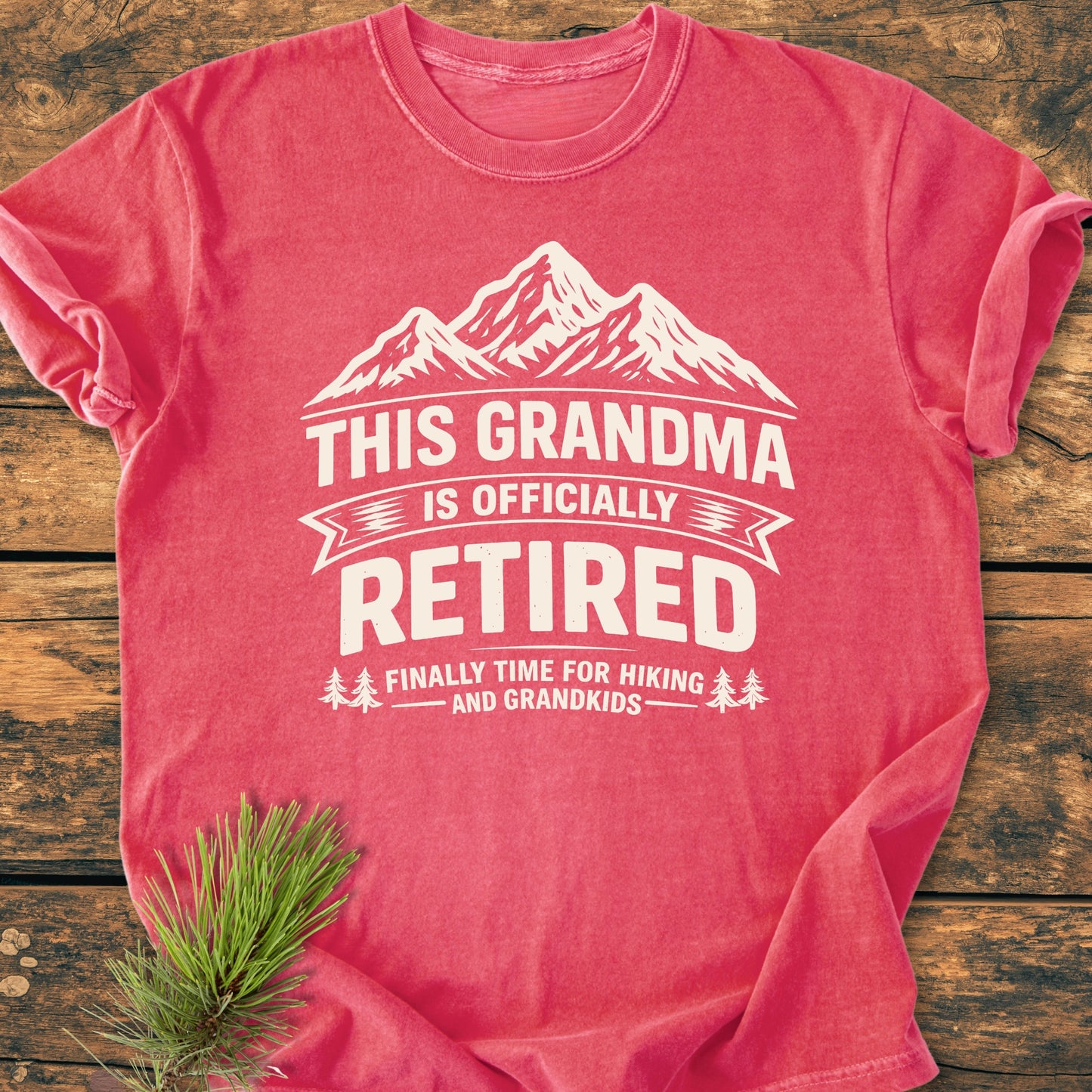 This Grandma is Officially Retired - Vintage Wash Tee features a white mountain graphic and retirement slogan, laid on wood with pine sprig, highlighting its cozy, ring-spun cotton fabric and durable construction.