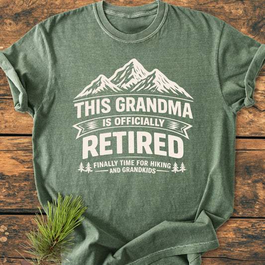 Heather-green This Grandma is Officially Retired - Vintage Wash Tee with mountain graphic and text, laid flat on wood. Features soft, garment-dyed fabric and relaxed fit, perfect for leisure activities.