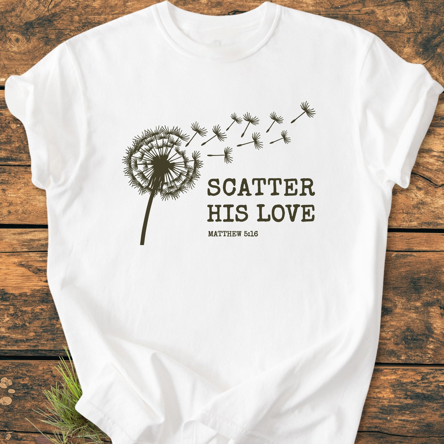 Scatter His Love, Matthew 5:16 - Vintage Wash Tee featuring a brown dandelion silhouette with drifting seeds and bold text on a soft, relaxed-fit white T-shirt laid on a wooden surface.