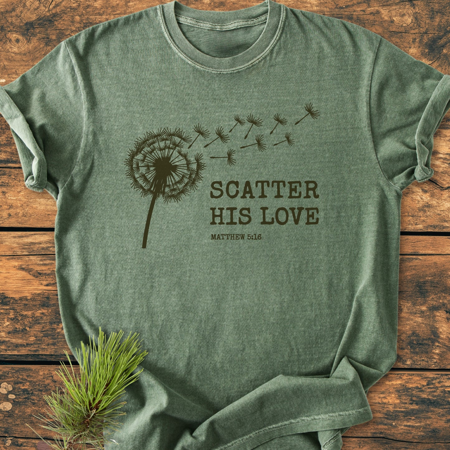 Scatter His Love, Matthew 5:16 - Vintage Wash Tee features a dandelion silhouette with seeds blowing away and text. Laid on wood, pine sprig beside, embodying a soft, relaxed fit for everyday wear.
