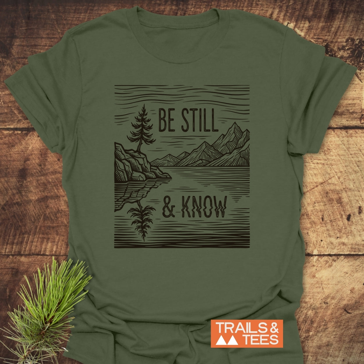 Be Still And Know T-Shirt in olive-green featuring a black line-drawing of a pine, mountain, and lake reflection, with 'TRAILS & TEES' logo, laid on weathered wood with pine sprig.