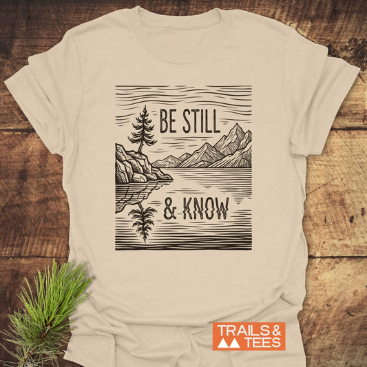 Be Still And Know T-Shirt featuring a woodcut-style tree illustration and reflective mountain lake, laid flat beside a pine branch, with an orange Trails & Tees logo in the corner.
