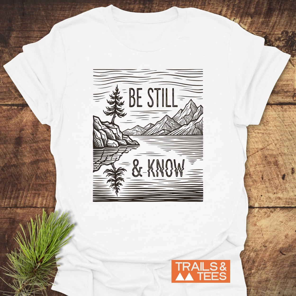 Be Still And Know T-Shirt featuring a mountain lake scene with a tree and peaks, accented by TRAILS & TEES logo, laid on wood with pine sprig nearby.