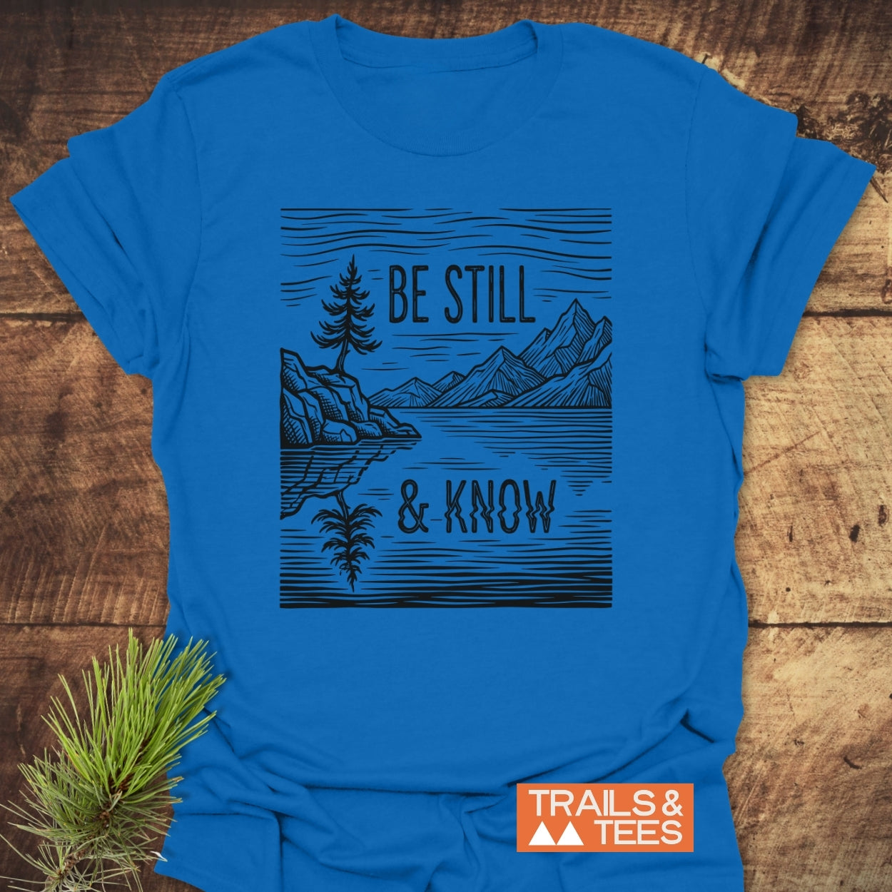 Be Still And Know T-Shirt featuring a wood-cut–style landscape with a conifer and mountains, printed on a soft, lightweight blue tee, ideal for nature-loving faith enthusiasts.