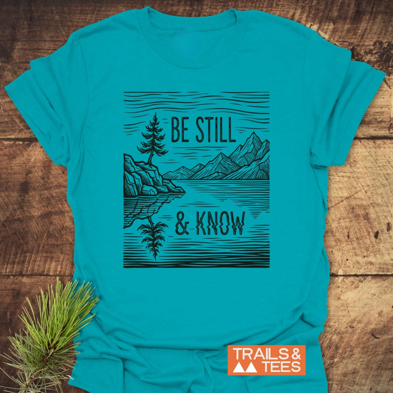 Teal Be Still And Know T-Shirt featuring a woodcut lakeside scene and TRAILS & TEES logo, styled with a pine sprig, laid on a wooden surface.