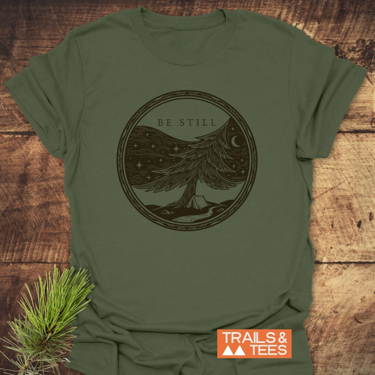 Be Still Softstyle T-Shirt featuring a stylized tree graphic with a tent and stream under a starry sky, highlighting tranquility. Labeled Trails & Tees in the corner.