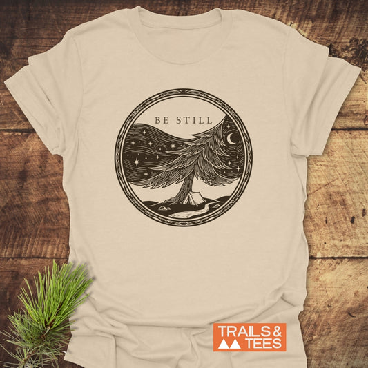 Be Still Softstyle T-Shirt features a woodcut-style illustration of a pine tree merging into a starry sky with a crescent moon, and a campsite at the base, conveying tranquility and faith.