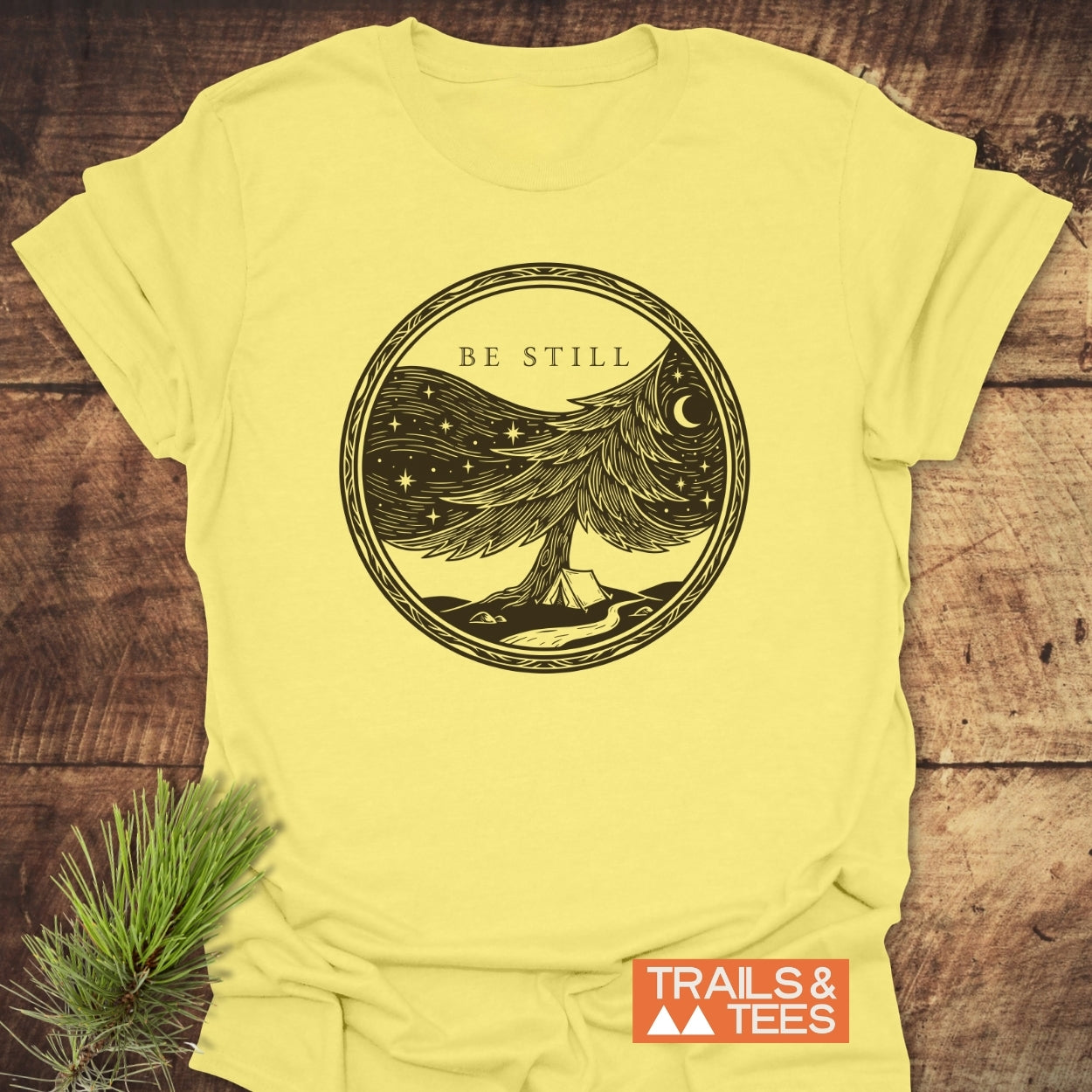 Be Still Softstyle T-Shirt featuring a windswept tree illustration with a night sky, campsite, and BE STILL text, laid flat with a pine sprig; ideal for nature-loving, faith-inspired individuals.
