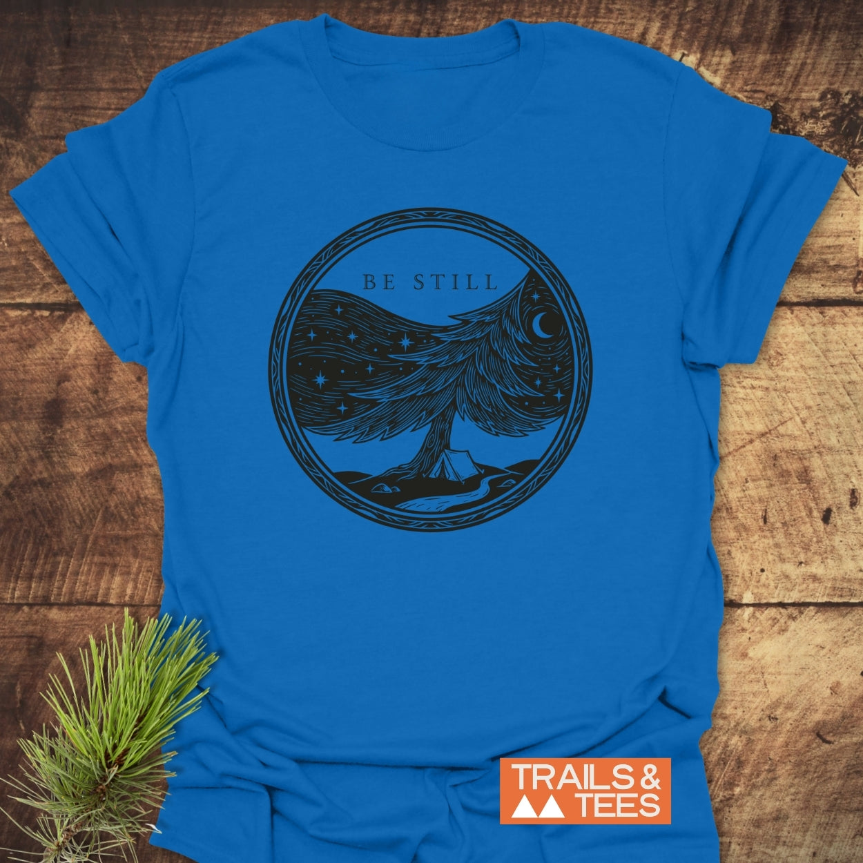 Be Still Softstyle T-Shirt featuring a hand-drawn pine tree graphic under a starry sky, with a crescent moon, tent, and stream, promoting a message of stillness and faith.