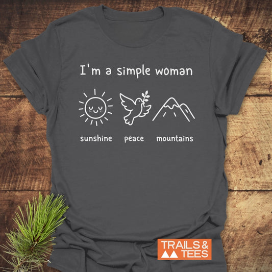 I'm A Simple Woman. Sunshine Peace Mountains T-Shirt featuring icons of a sun, dove, and mountains, laid on wood with pine, conveying serenity and love for nature by Trails & Tees.