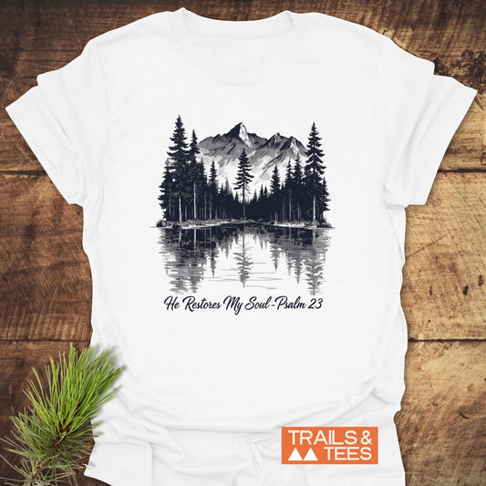 He Restores My Soul T-Shirt with a sketch of pine trees, mountains, and lake reflection. Includes the text “He Restores My Soul – Psalm 23,” accompanied by a small pine branch and orange “TRAILS & TEES” logo.