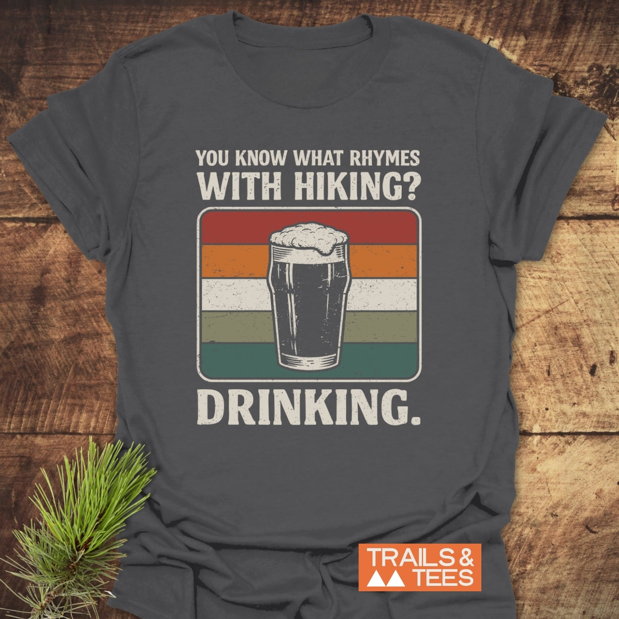 What Rhymes With Hiking? Softstyle T-Shirt featuring a beer glass design with vintage stripes, laid on wood with a pine sprig; Trails & Tees logo visible.